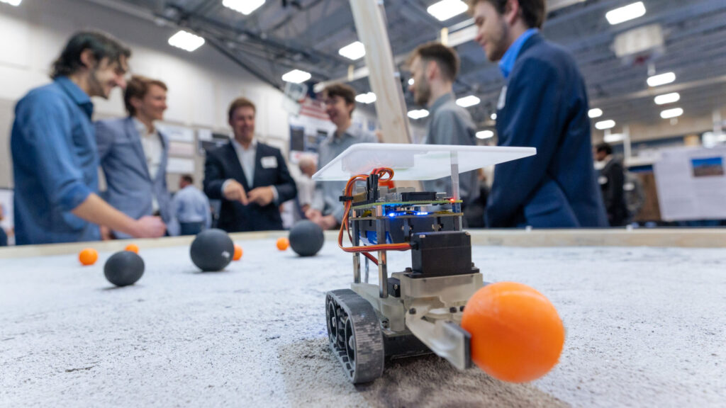 forefront of image shows a miniture robot holding a ping pong ball with group of students presenting in background