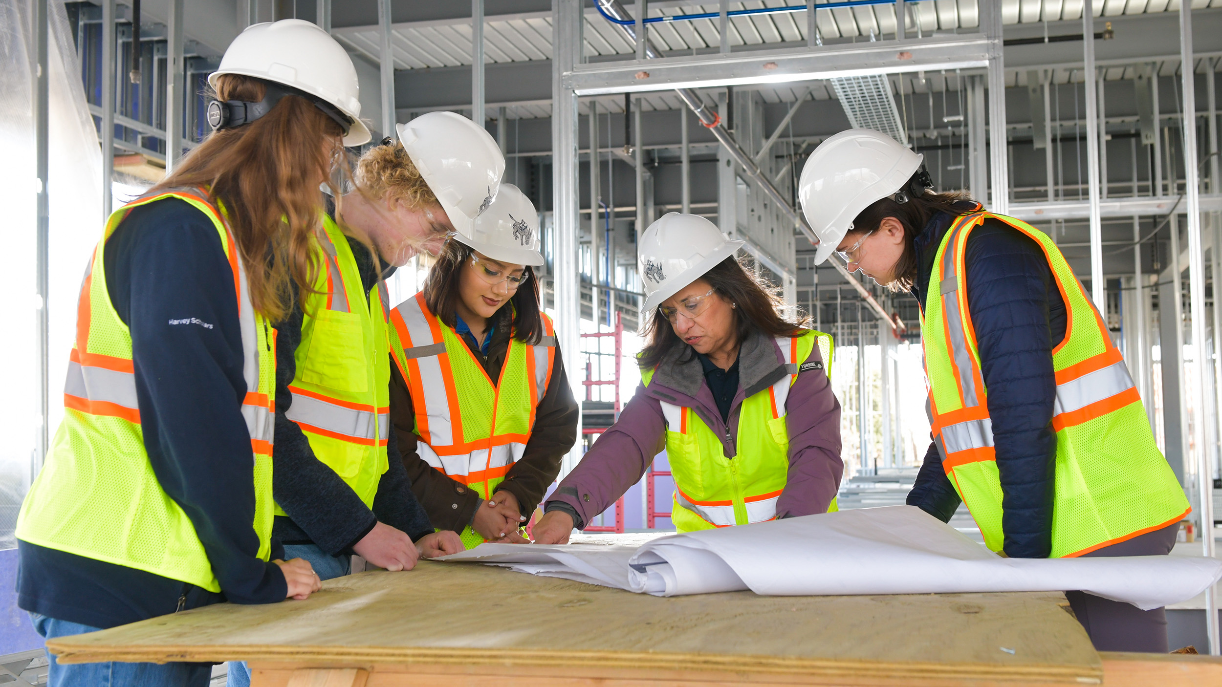 Five people wearing white hard hats, reflective safety vests, and safety glasses, standing around a table with construction plans or blueprints.