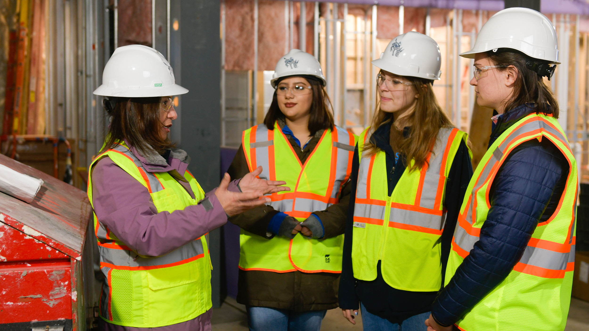 Four people on a construction site, all wearing white hard hats, reflective safety vests, and safety glasses.