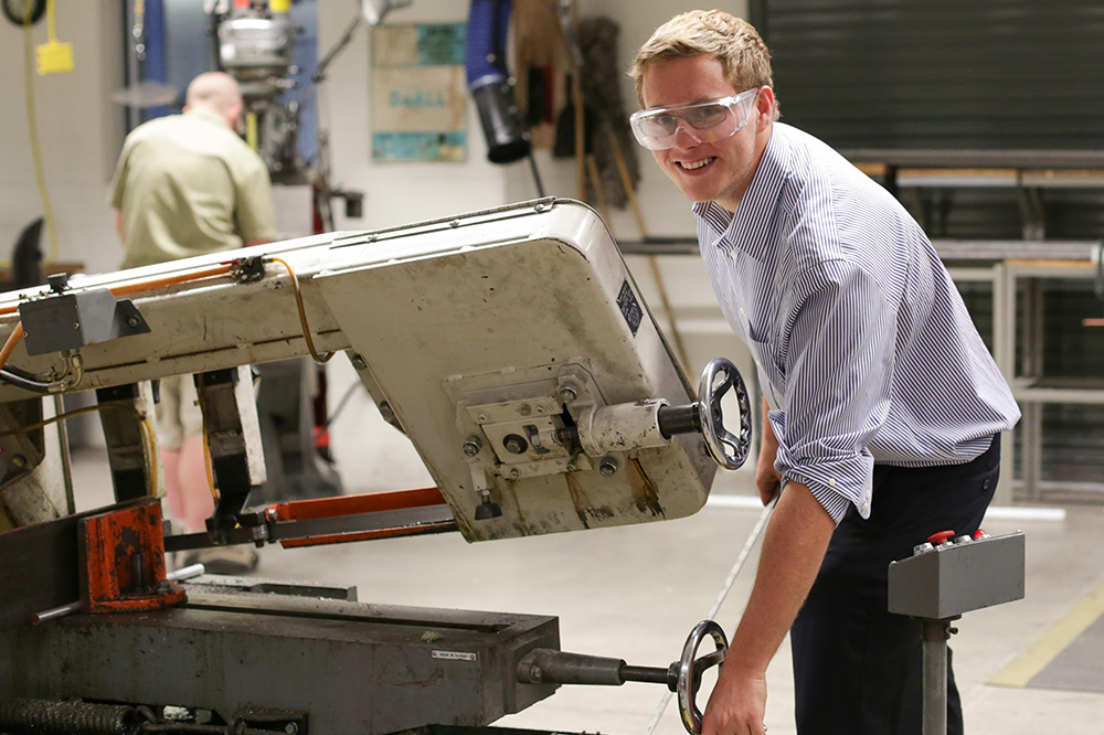 A man turning a wheel on a machine, leaning over, wearing safety goggles and a blue striped shirt